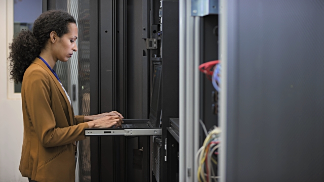 Medium locked down shot of a female technician analyzing server functions in the server room using a laptop.